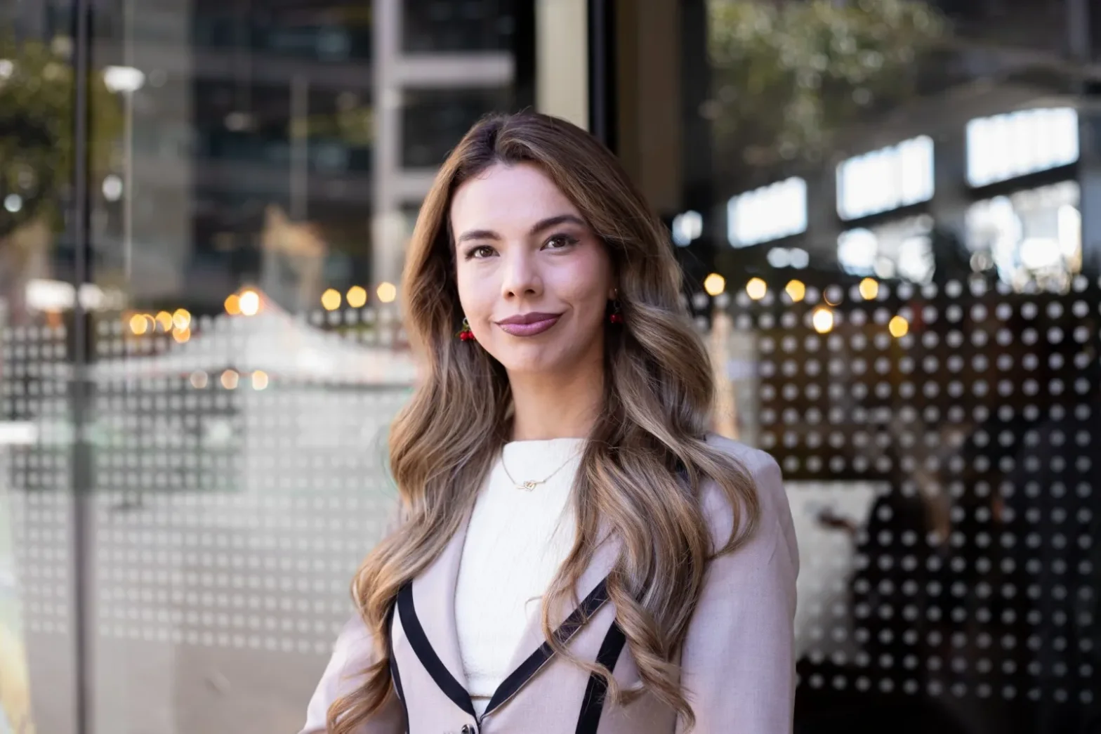 Portrait of a smiling woman with long wavy hair in a light coat, standing outside a glass building with blurred city lights behind her.