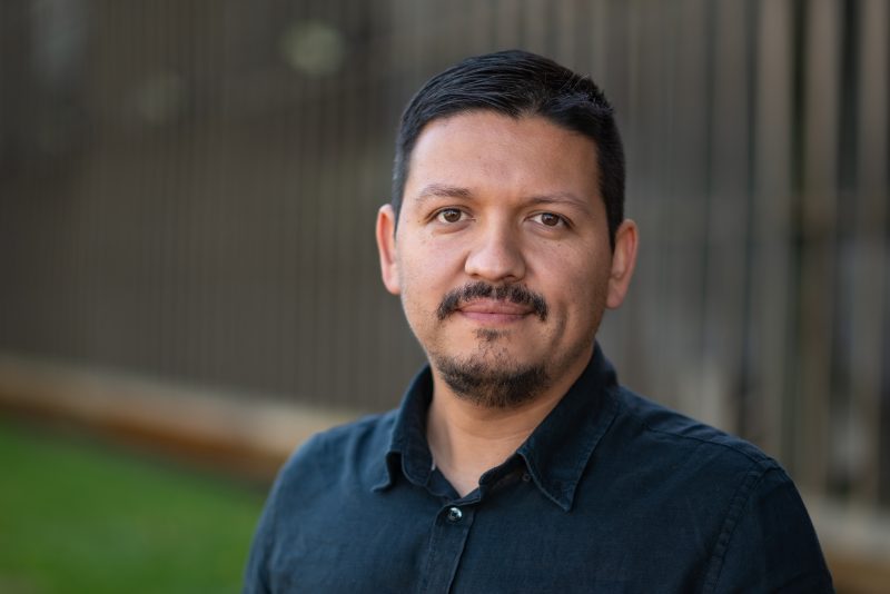 Portrait of a man with short dark hair and a goatee, wearing a navy shirt, looking at the camera outdoors.