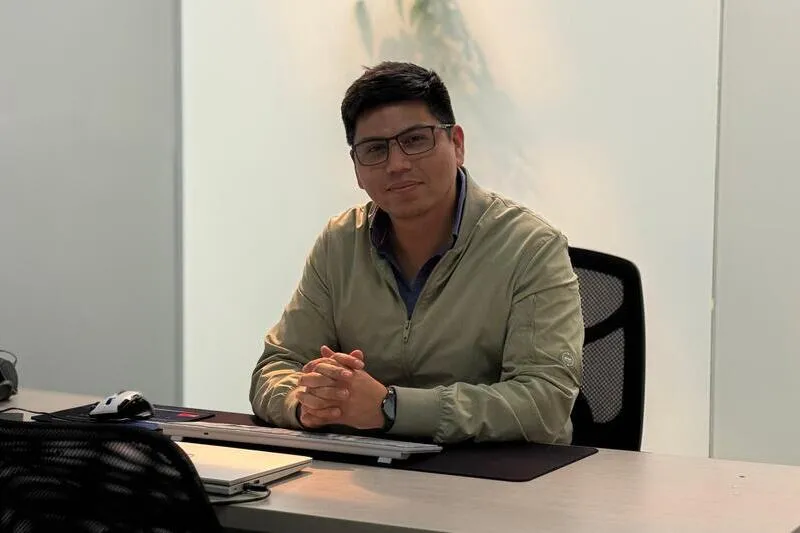 Man wearing glasses and a beige jacket sits at a desk in an office, hands folded, with a laptop in front.