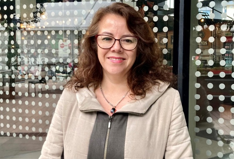 Smiling woman with curly brown hair and red glasses standing outside a storefront with dotted glass panels behind her.