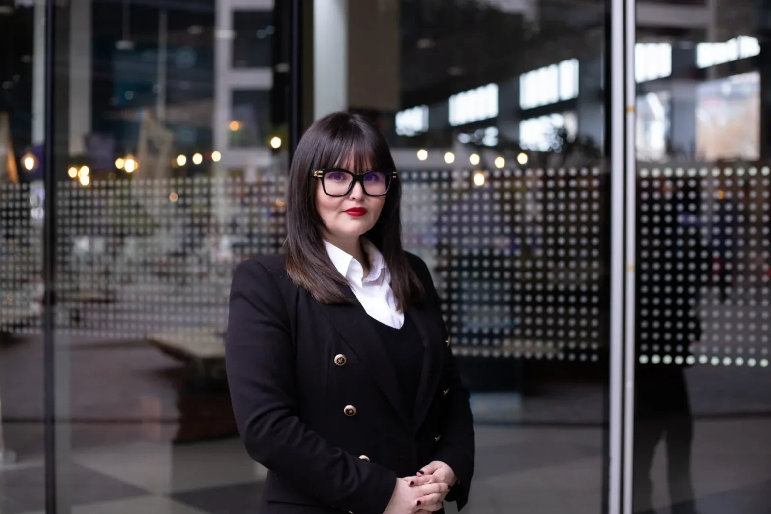 Professional woman in a black blazer and white shirt stands before glass doors with a dotted pattern.