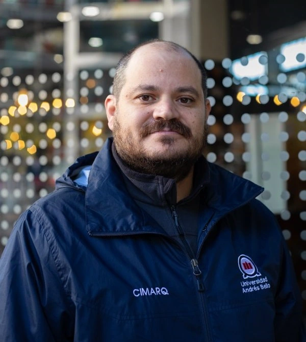Portrait of a man with a beard wearing a dark blue jacket with CIMARQ and Universidad Andrés Bello logos, standing in front of a glass wall.