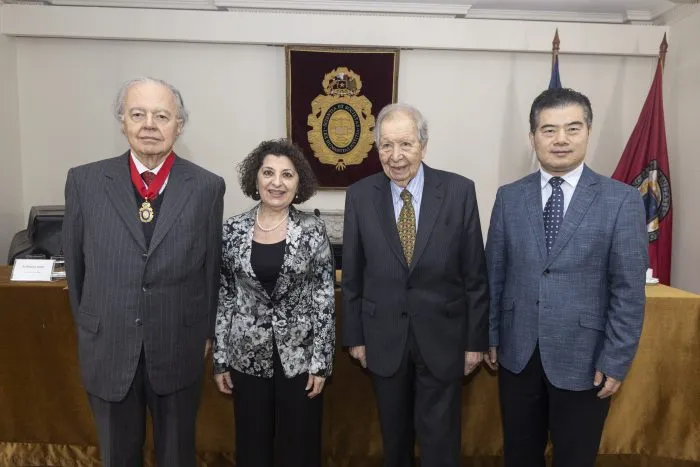Four formally dressed individuals stand side by side in a conference room, with a crest on the wall behind them, suggesting an award or ceremony.