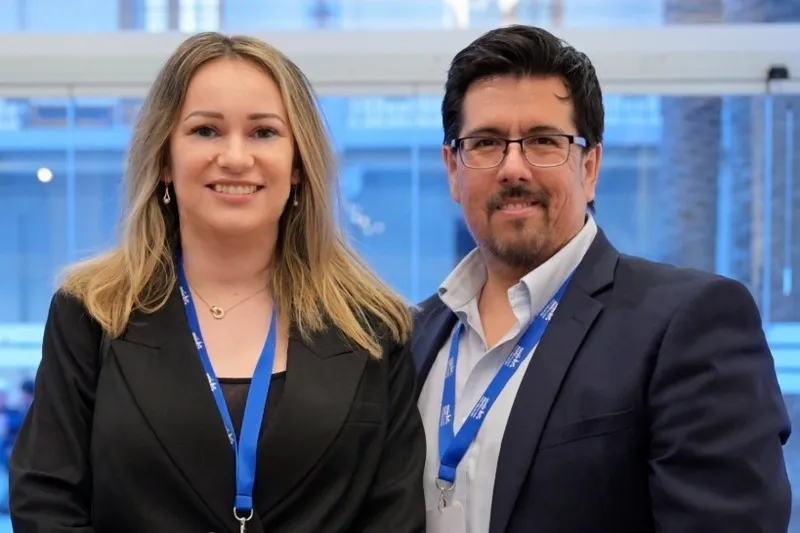 Two professionals pose for a photo at a conference, wearing business attire and blue lanyards with badges visible in a bright modern venue.