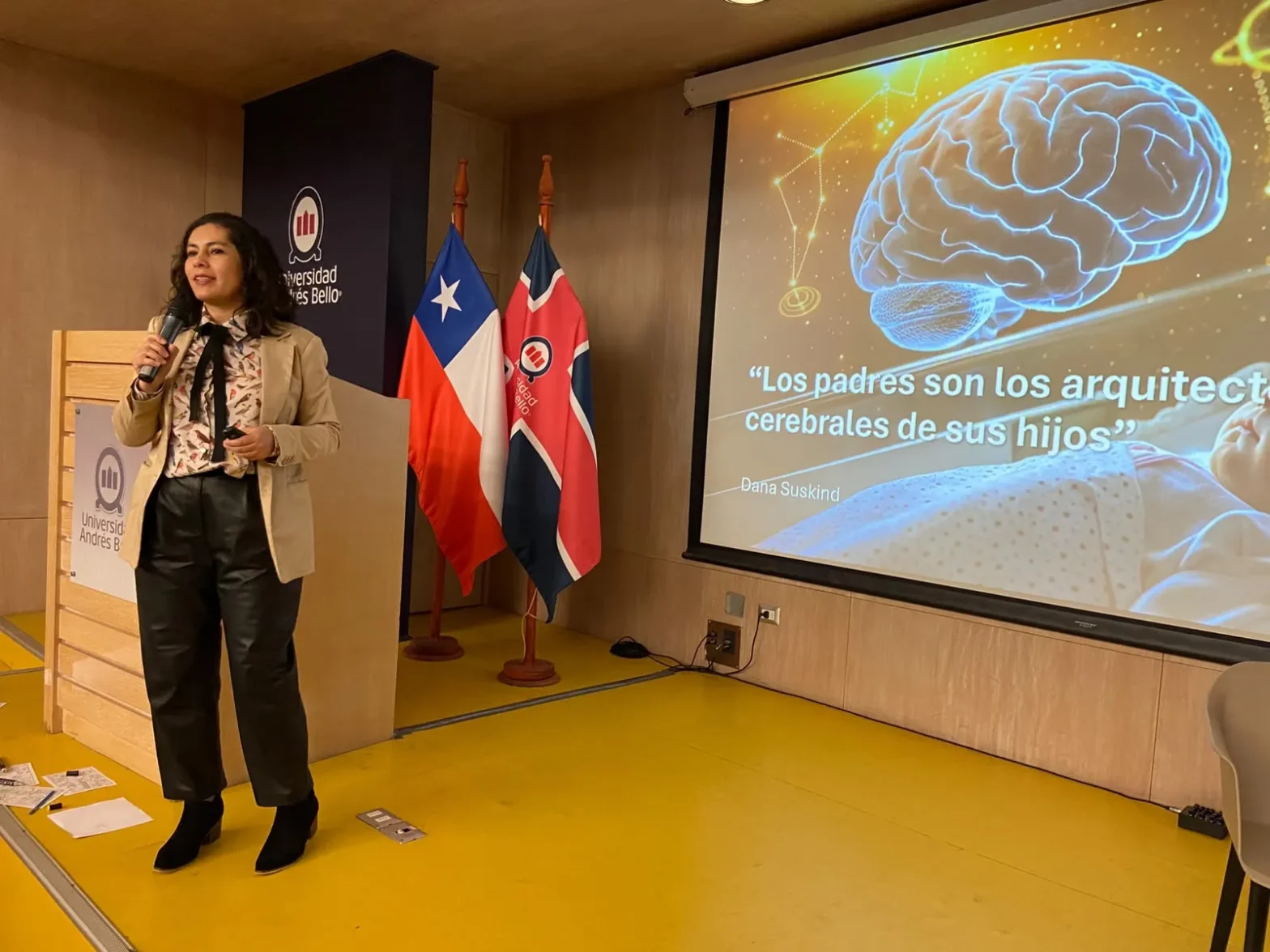 Woman speaker at a podium giving a lecture; a large brain-themed slide with a Spanish title is projected beside her.