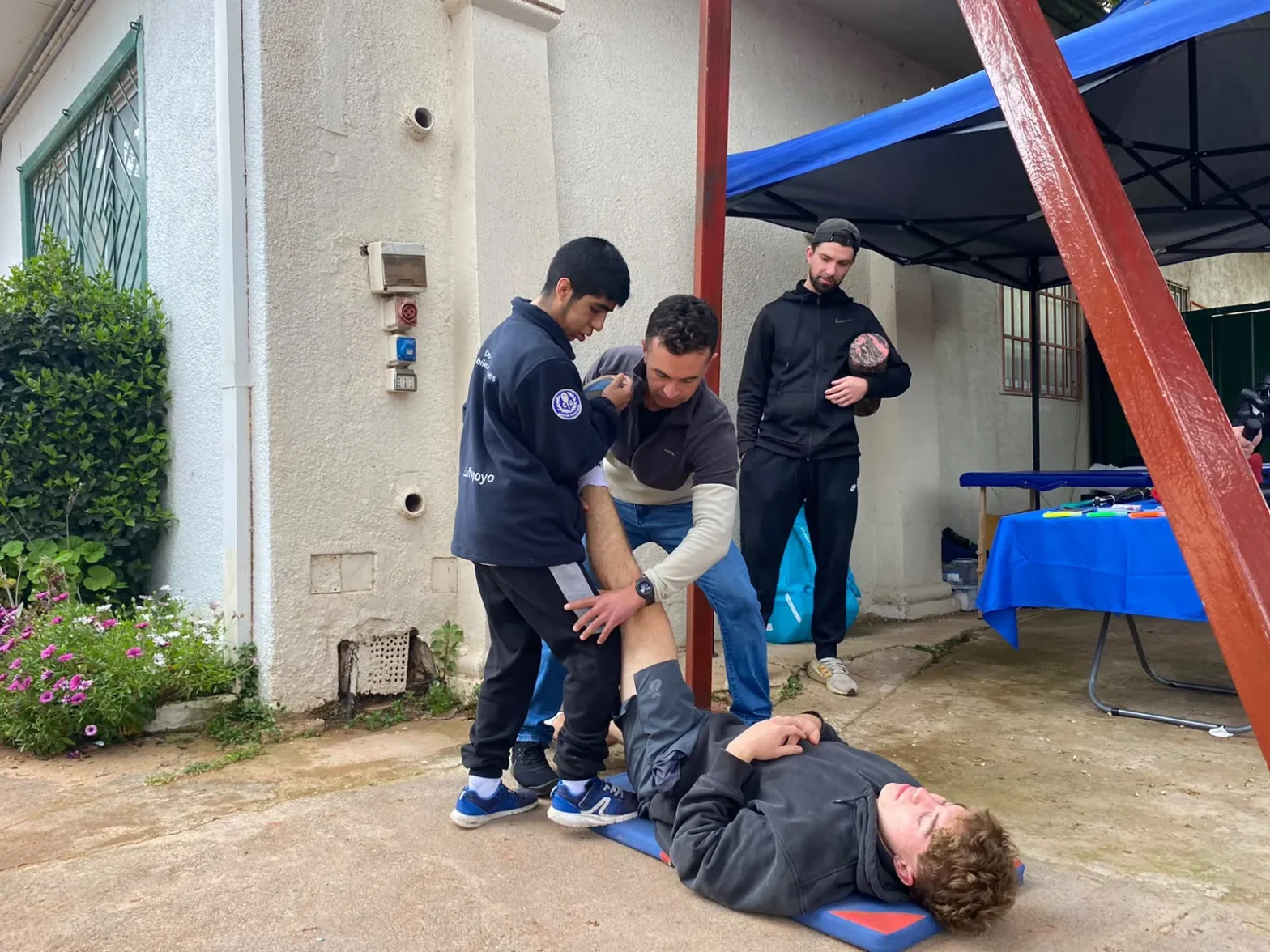 Two trainees help a person lying on a mat outdoors during a first aid training session under a blue canopy, as another observer watches nearby.