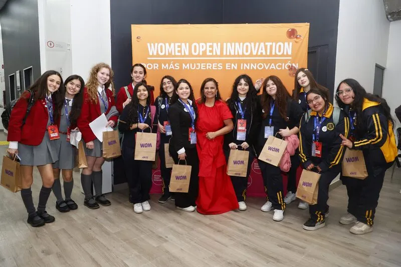 Group of young women posing at the Women Open Innovation event in front of an orange banner; many wear lanyards and hold gift bags.