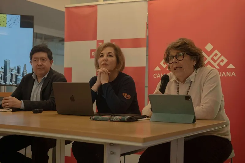 Panel discussion with three speakers at a table, laptops and a tablet in front, red banners behind them.