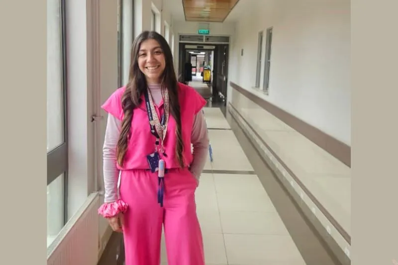 Young woman wearing bright pink medical scrubs stands in a hospital hallway, smiling, with a lanyard and ID badge nearby in the frame.