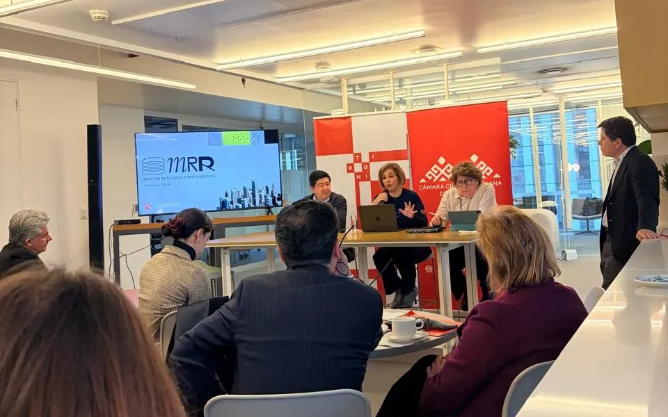 Panel discussion in a bright office: three people seated at a table with laptops, speaking to an audience while a screen shows 'MRR' or similar branding and red banners in the background.