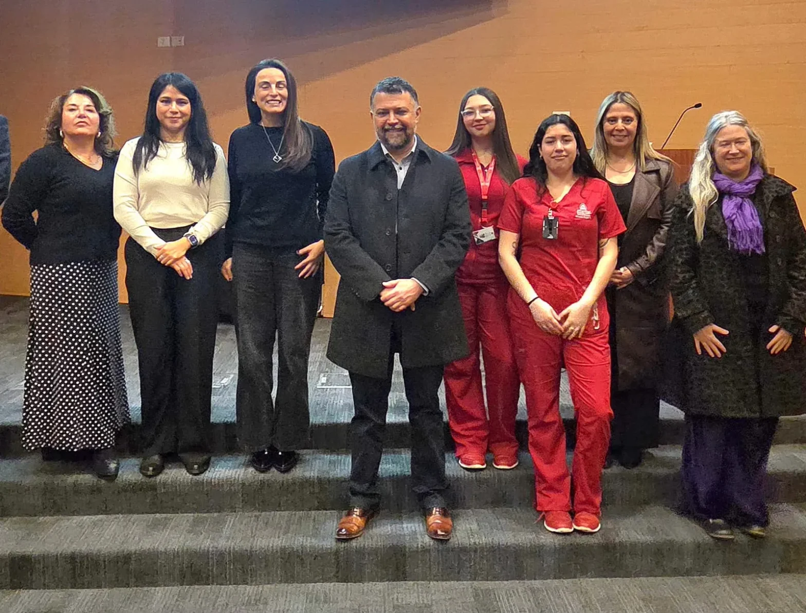 Eight people standing on a stage in front of a wooden wall, posing for a group photo; mix of business attire and red medical scrubs.