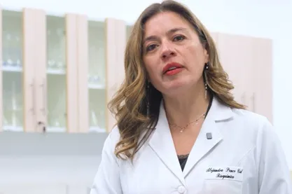 Female medical professional in a white lab coat speaking in a clinical setting, with cabinets in the background.