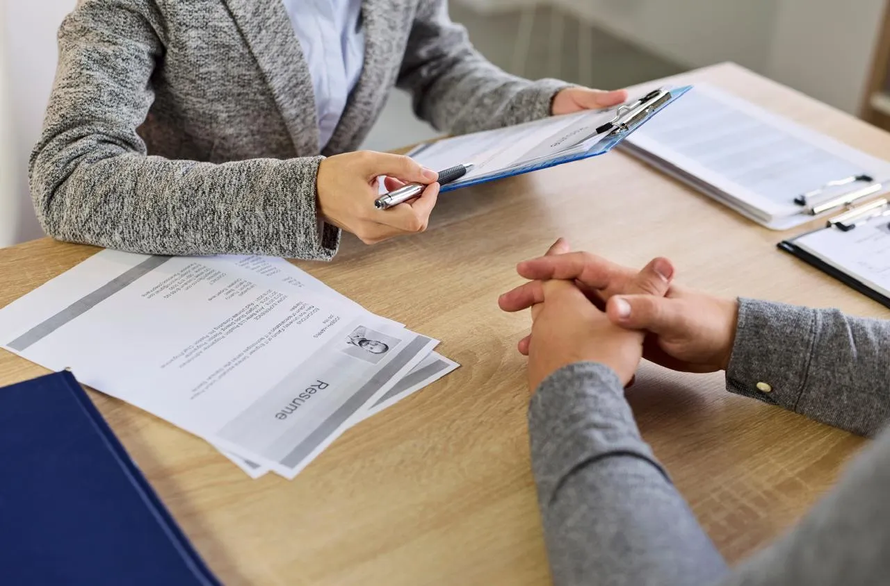 HR interviewer reviewing a resume on a clipboard while a candidate sits with hands folded on the table, documents and a resume on the desk nearby