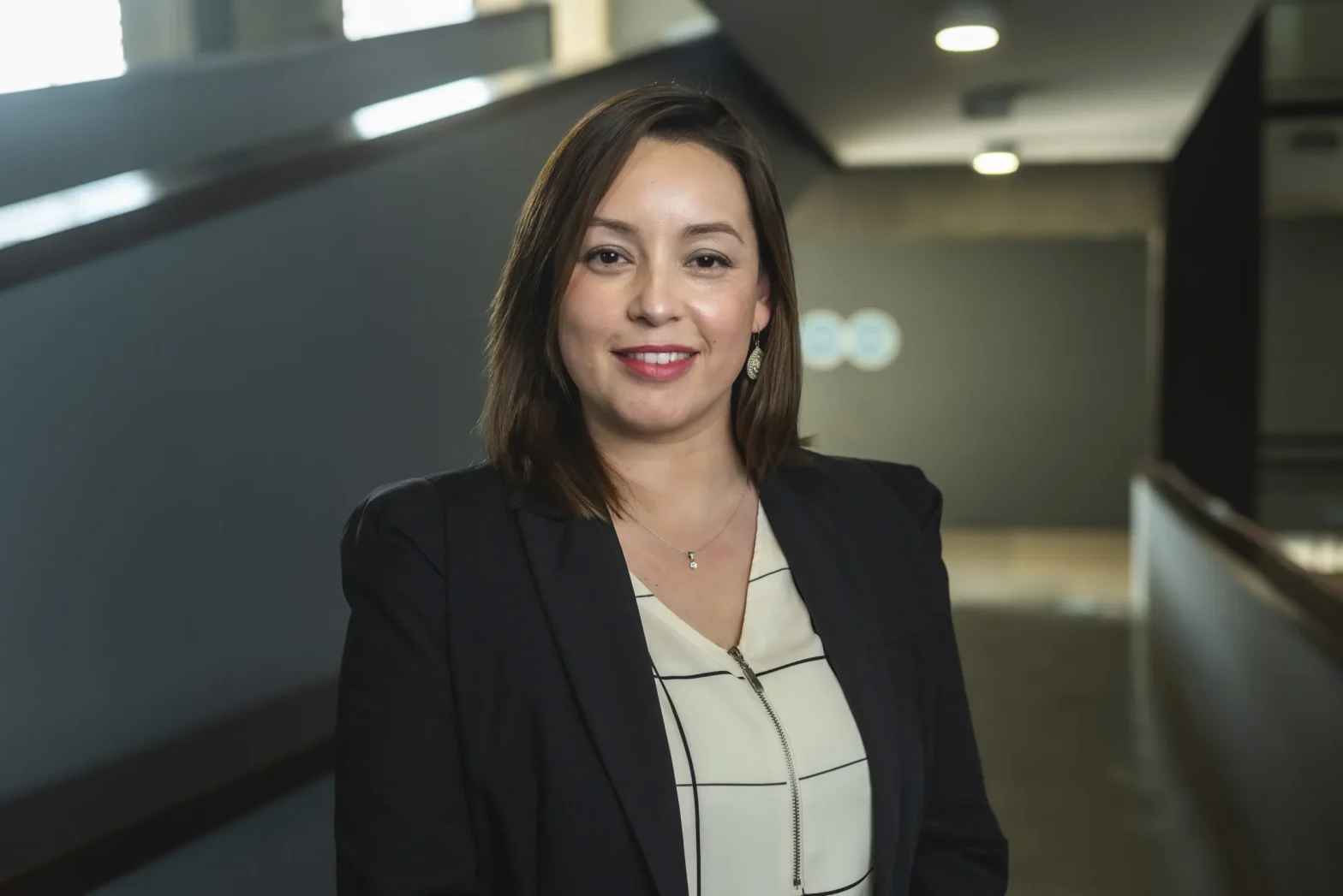 Smiling professional woman in a black blazer standing in a modern office hallway in the background.