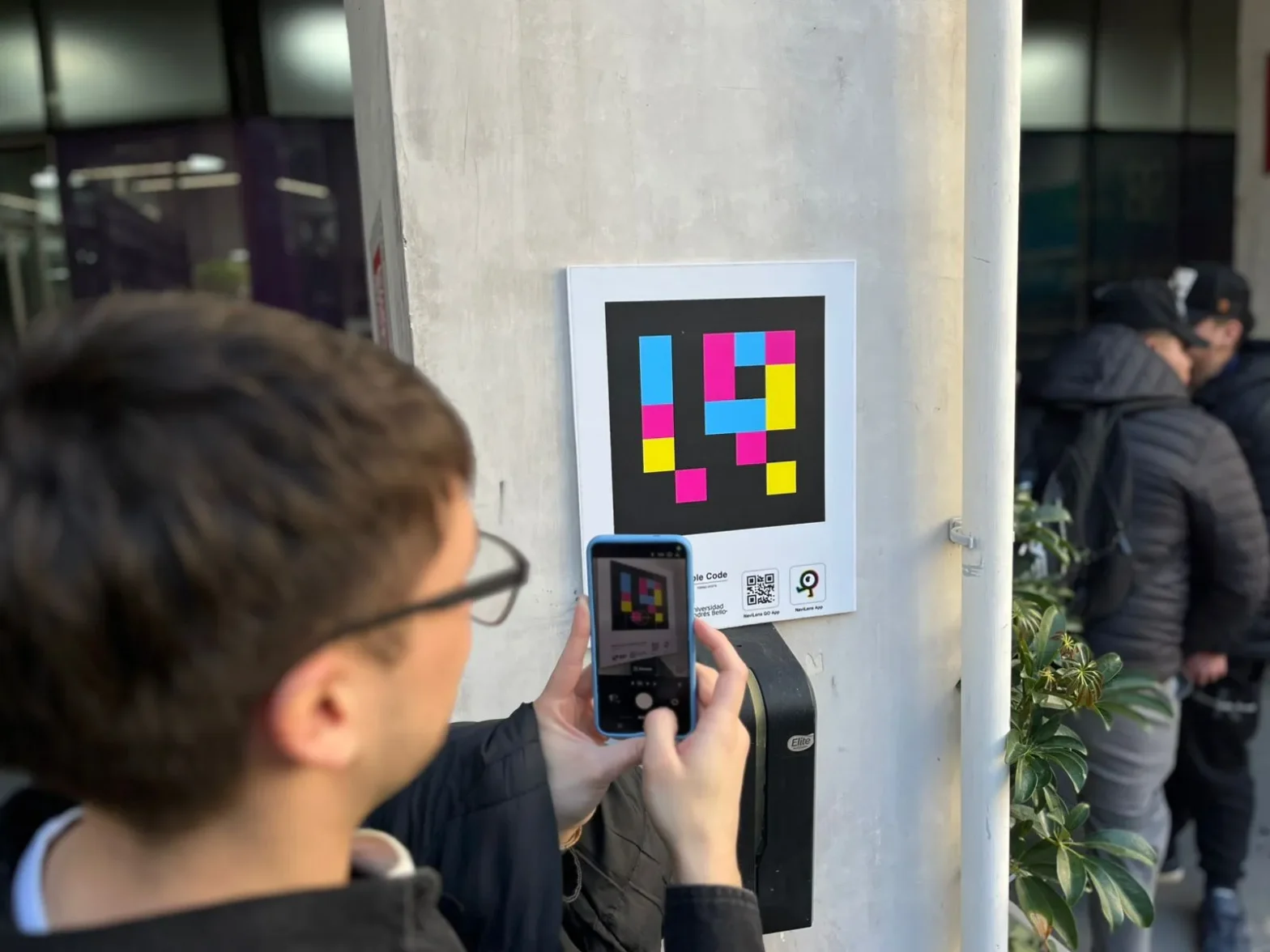 Person taking a photo of a colorful pixel-art poster mounted on a light-gray pillar outside.