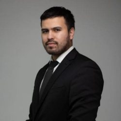 Professional headshot of a man in a black suit and gray tie, facing slightly left against a neutral gray background.